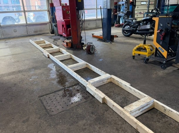 Long white metal frame or ladder positioned on wet concrete floor in an automotive garage workshop with various equipment and tools visible