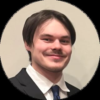 Professional headshot of a smiling man with dark hair wearing a black blazer, white dress shirt, and dark tie against a beige background