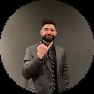 Professional headshot of a man with beard wearing gray blazer and striped tie, posing with hand near chin against neutral background